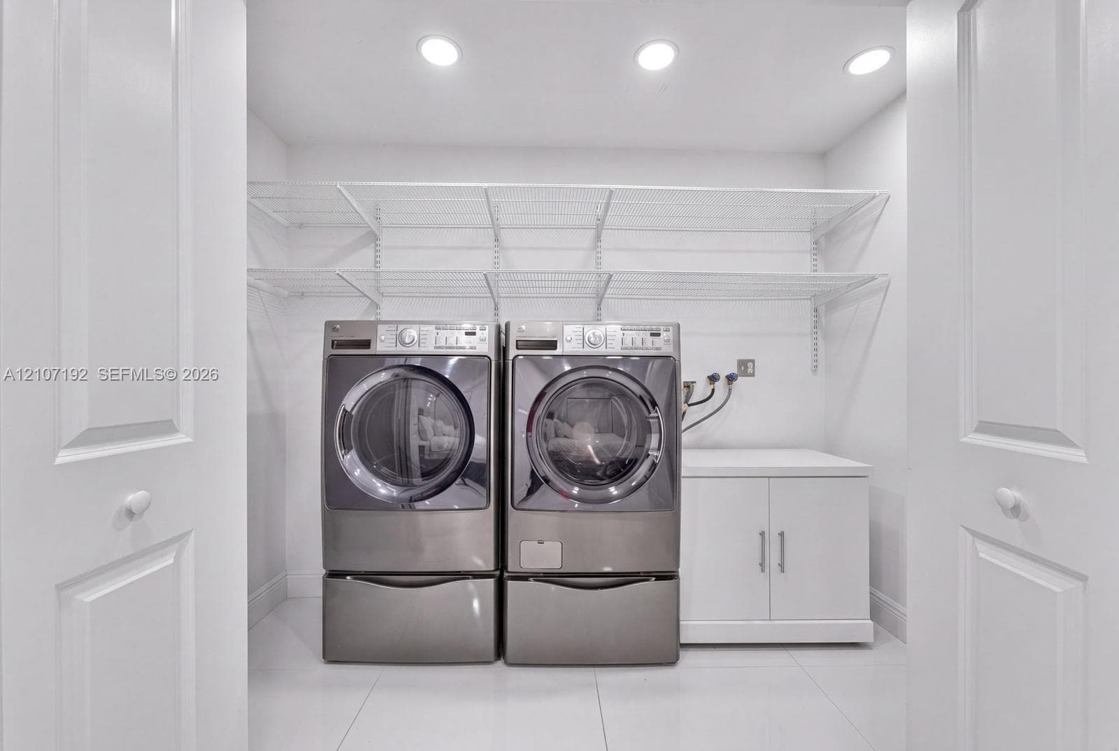 Laundry area featuring large-format tile flooring, recessed lighting, white base cabinetry with dual doors, tiered wire shelving, and double-door entry
