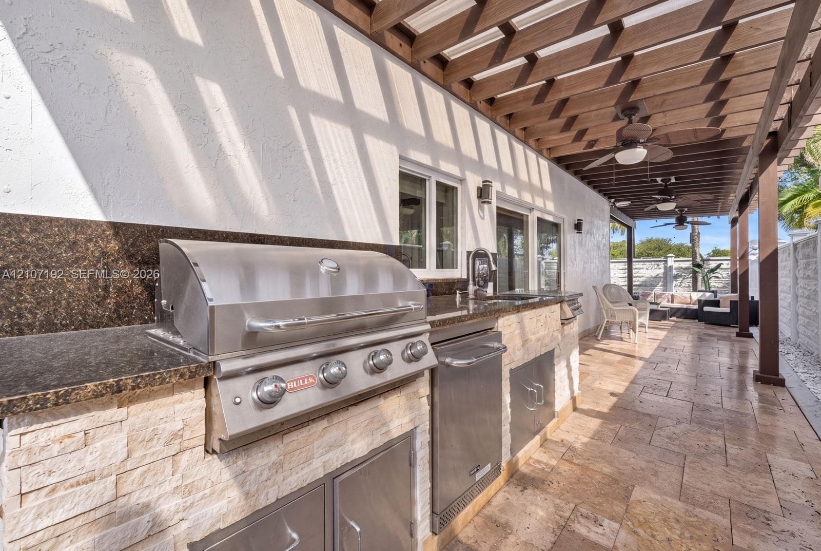 Outdoor kitchen featuring a stone-clad base, dark countertop, built-in stainless steel grill, stainless steel under-counter storage, and a wooden pergola cover over stone-tiled patio flooring