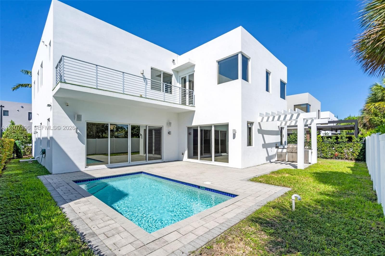 Modern two-story exterior featuring smooth white stucco siding, second-level balcony with horizontal metal railing, in-ground swimming pool, stone-paver patio, and pergola-covered outdoor cooking station