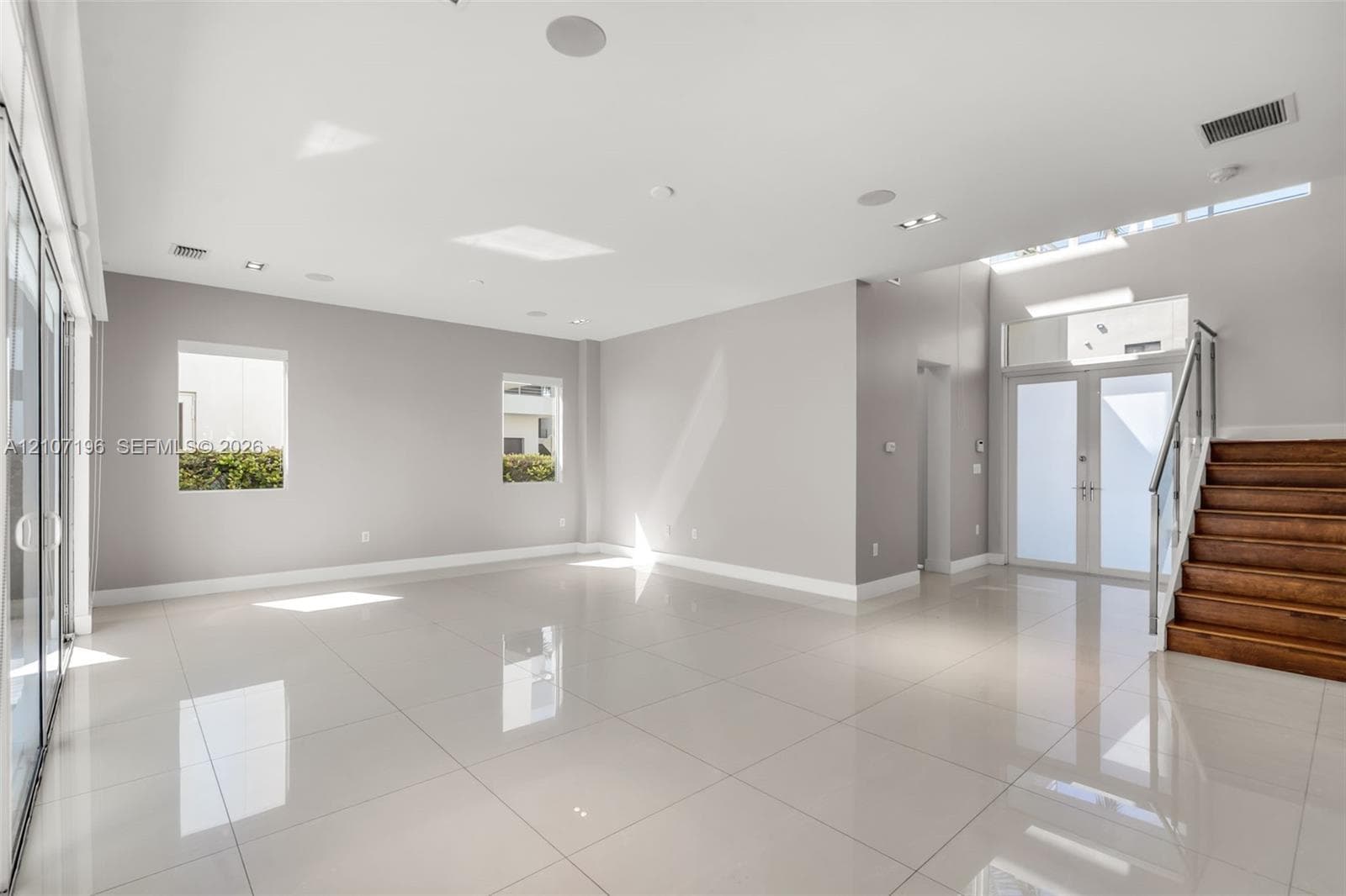 Open-concept interior featuring large-format tile flooring, recessed ceiling lighting, a wood-tread staircase with glass railing, and oversized glass entry doors