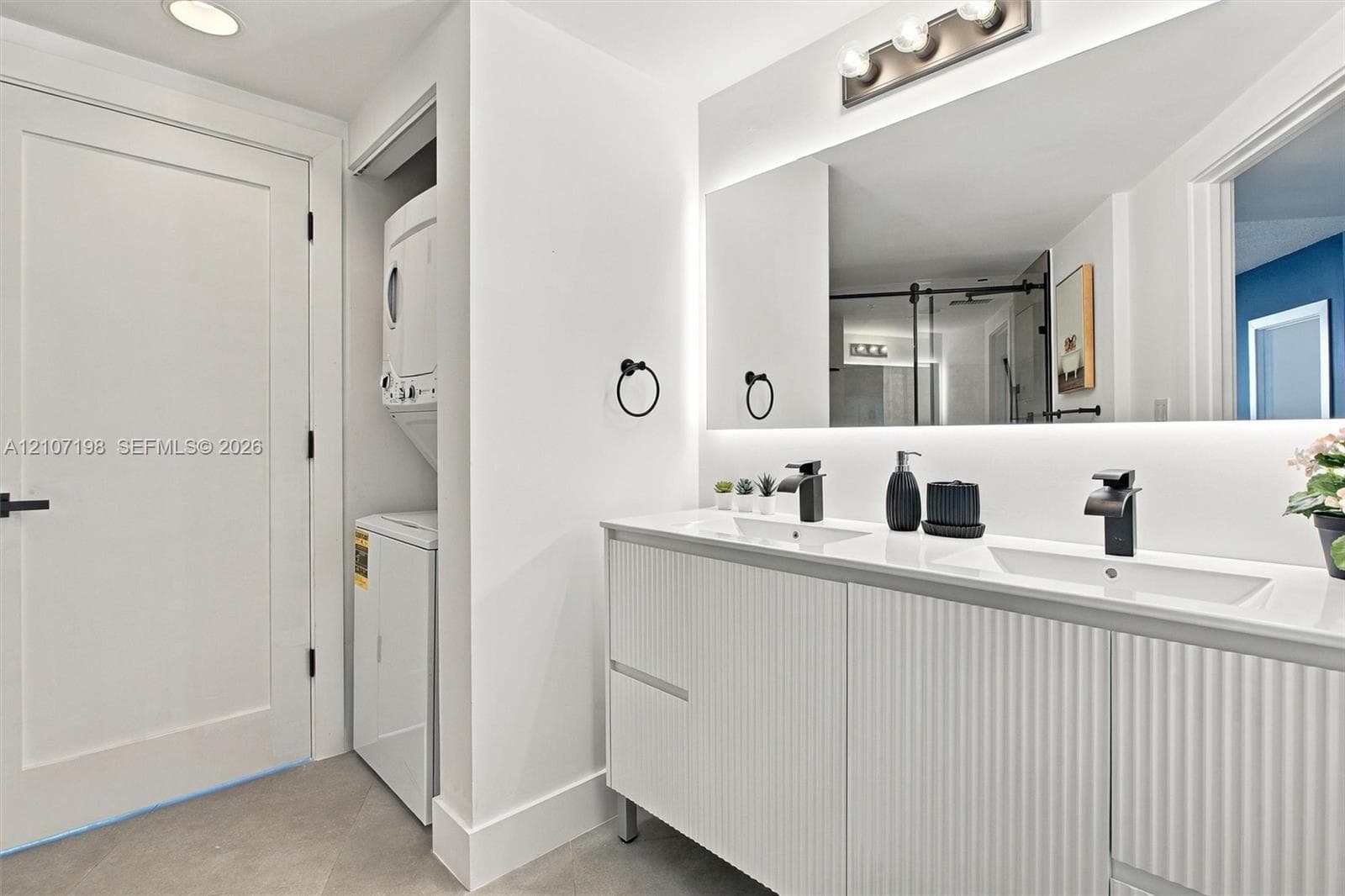 Double-vanity cabinet with fluted detailing, integrated solid-surface countertop, matte black fixtures, full-width mirror with backlighting, and built-in laundry station
