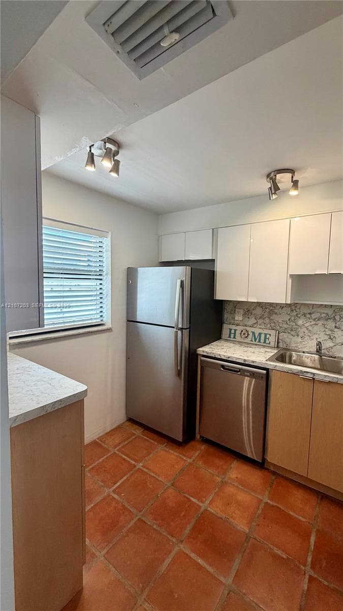 Kitchen featuring terracotta tile flooring, stainless steel appliances, light-toned cabinetry, stone-patterned countertops, and dual track lighting fixtures