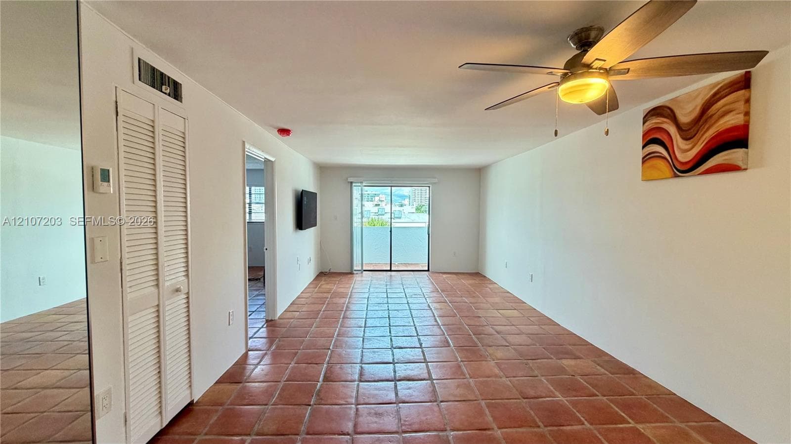 Large open-concept room featuring terra cotta tile flooring, floor-to-ceiling mirror paneling, a ceiling fan with integrated lighting, and sliding glass doors
