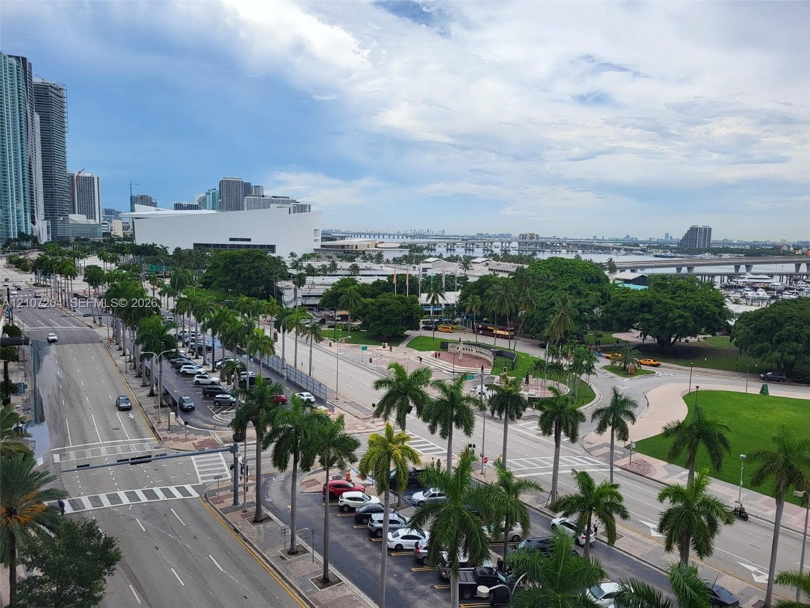 High-level urban perspective showcasing metropolitan skyline, extensive paved thoroughfares, mature palm tree-lined landscaping, waterfront proximity, and public park grounds