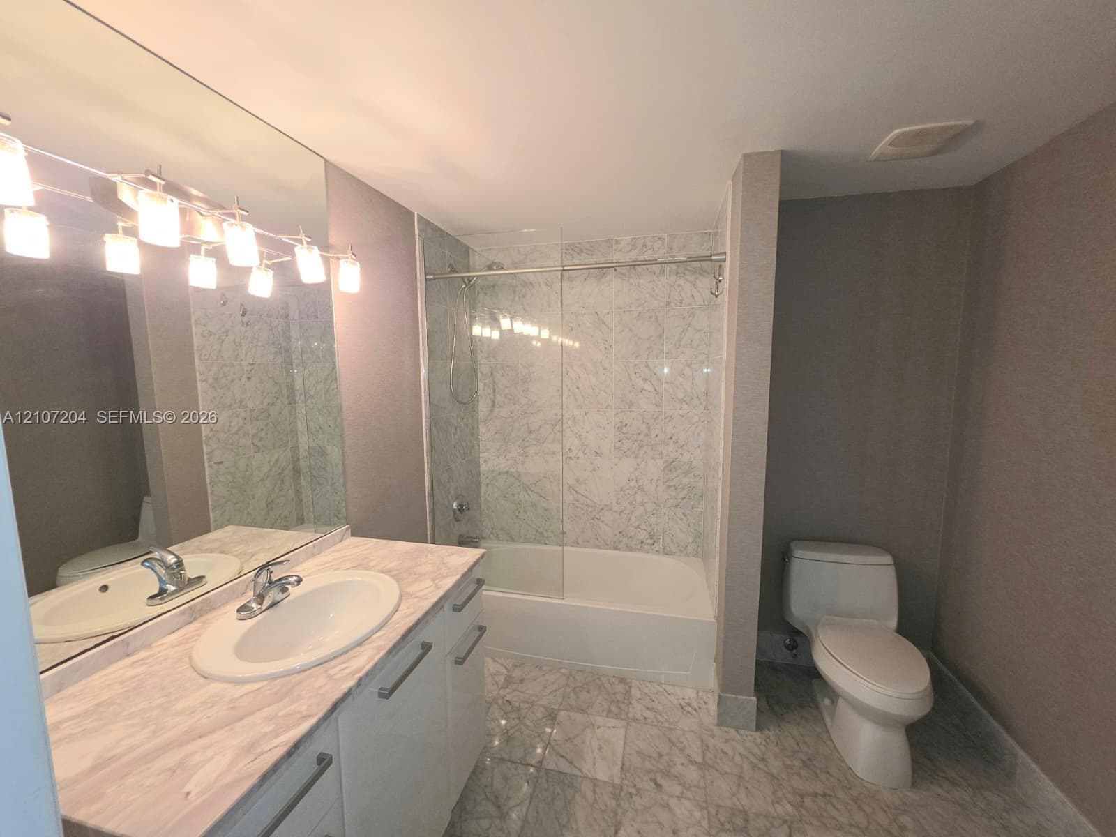 Bathroom featuring a vanity with stone-finish countertop and integrated basin, full-width wall mirror, chrome-finish multi-light fixture, stone-tile flooring, and a shower-tub combination with glass enclosure and stone-tile surround