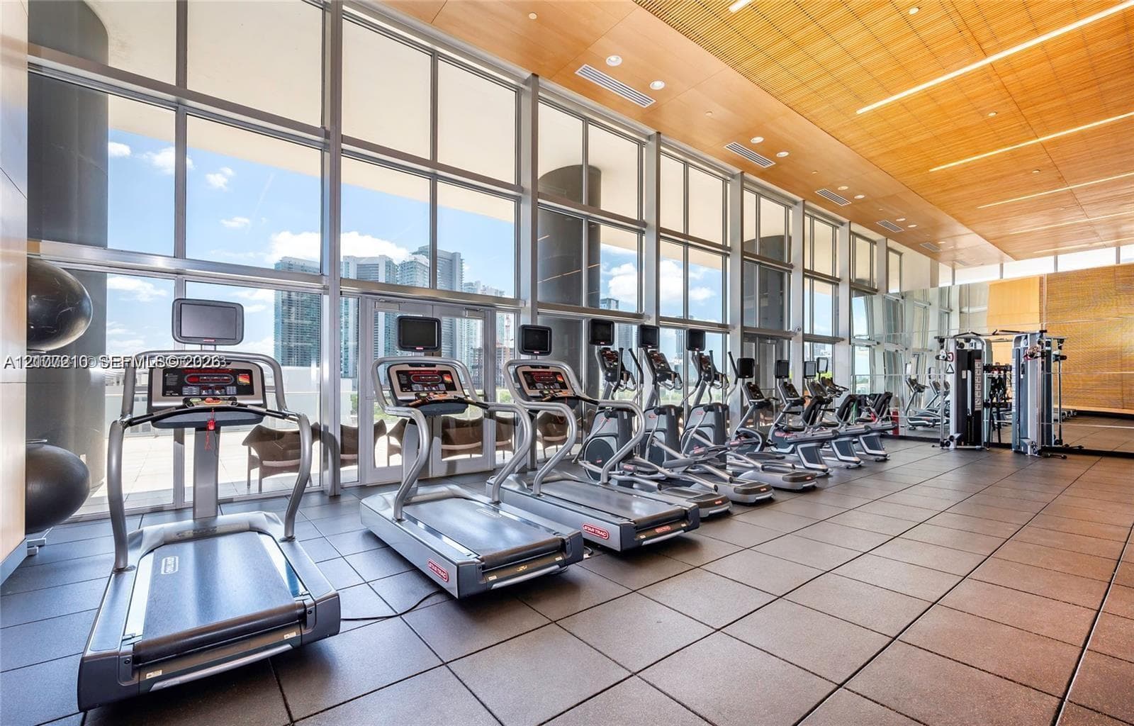 State-of-the-art fitness center featuring an expansive wall of floor-to-ceiling windows with skyline vistas, polished tile flooring, and a comprehensive array of cardio and strength training equipment
