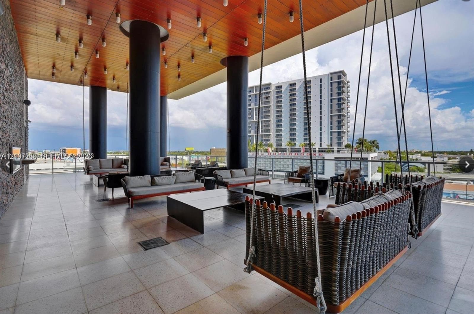 Covered outdoor lounge featuring a wood-panel ceiling with recessed lighting, prominent black structural columns, a textured stone feature wall, and large-format floor tiling