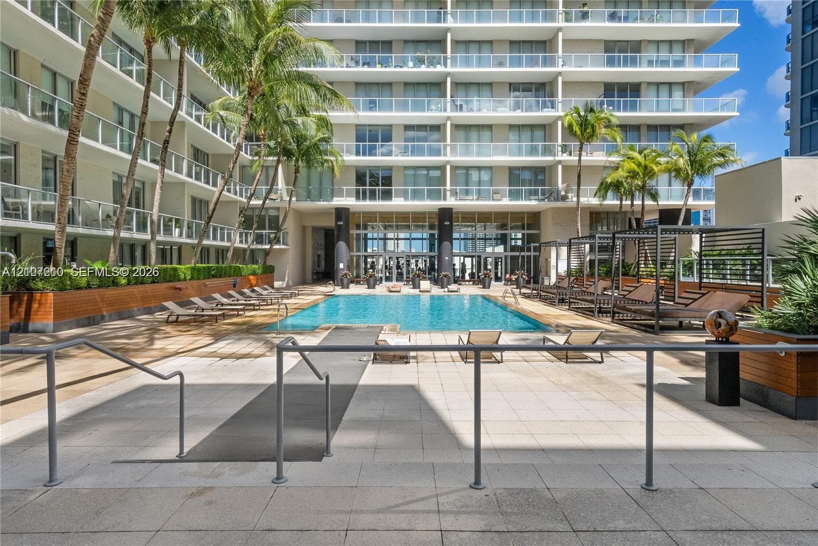 Swimming pool with surrounding paver deck, palm tree landscaping, and multi-story building exterior featuring glass-paneled balconies
