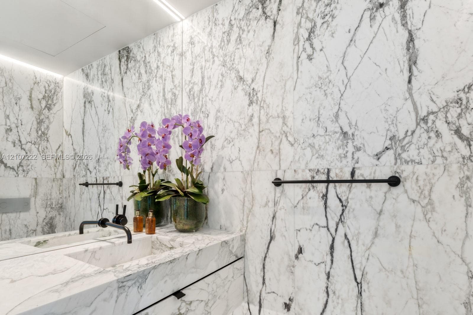 Full-height stone wall cladding featuring integrated cove lighting, vanity with matching stone countertop, undermount sink, and matte black wall-mounted fixtures