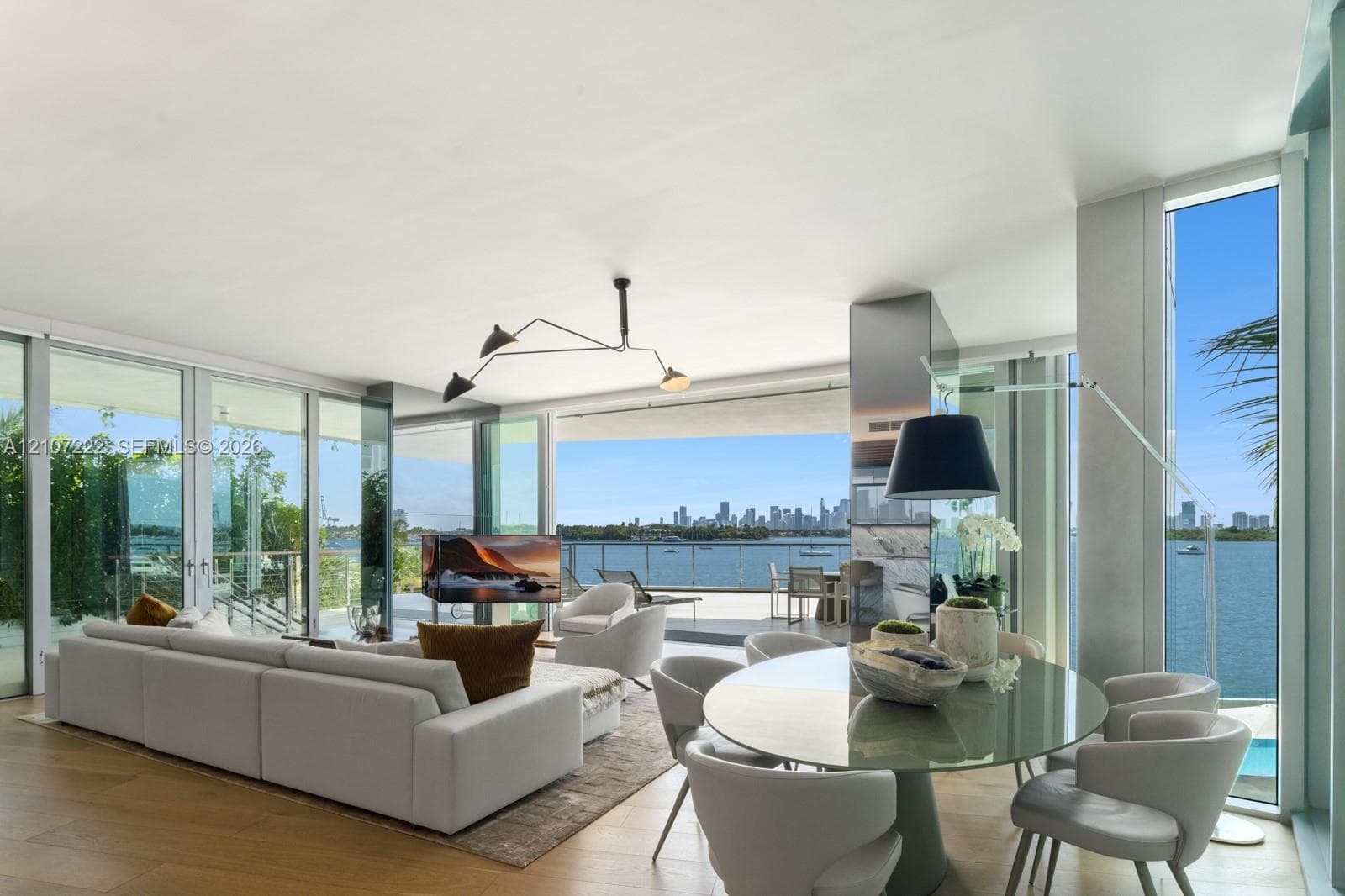 Floor-to-ceiling glass walls, wood-finish flooring, recessed architectural lighting, and a large private balcony with water and city skyline scenery