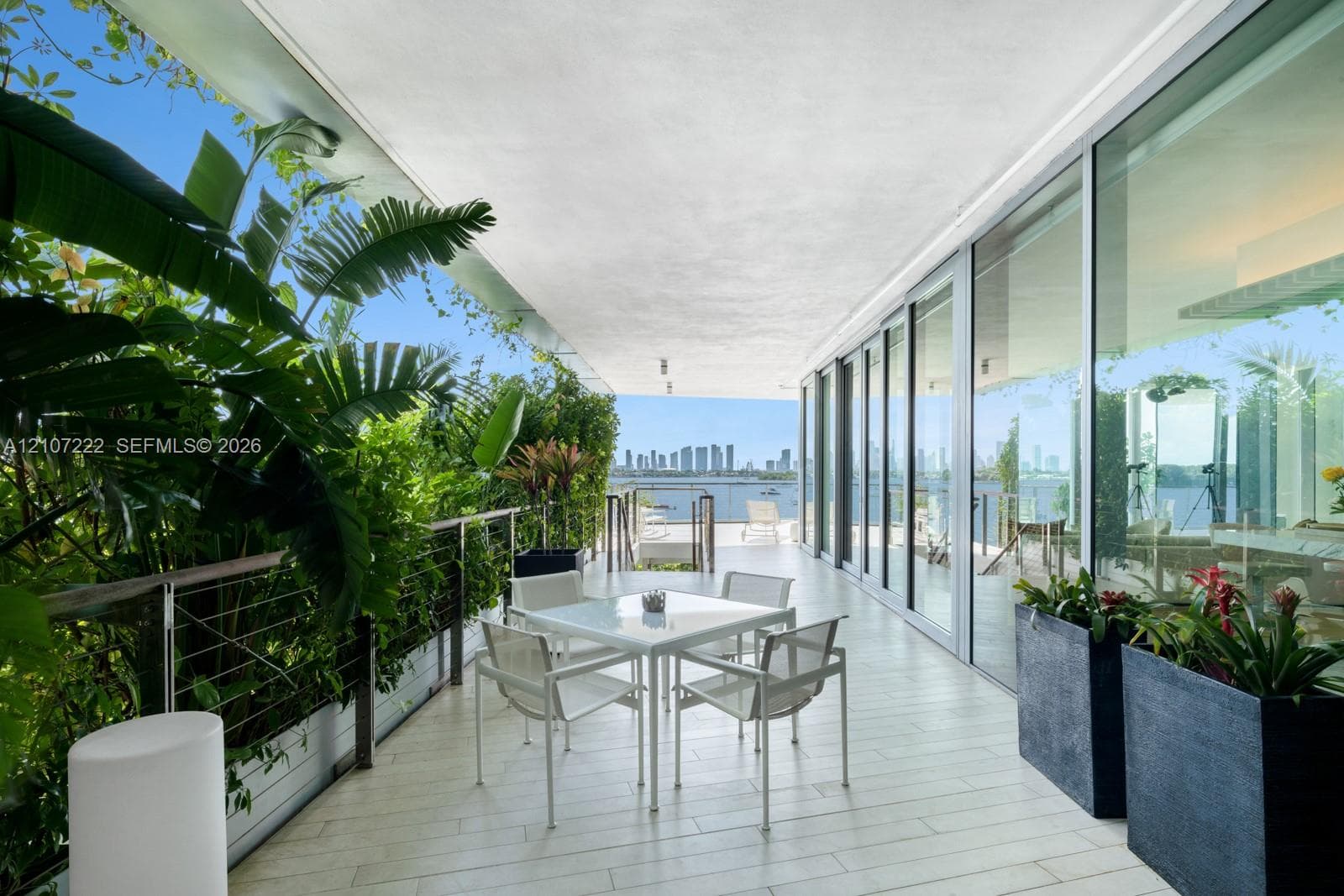 Covered outdoor terrace featuring light-toned tile flooring, floor-to-ceiling glass panel walls, a stainless steel cable railing system, and integrated built-in planters