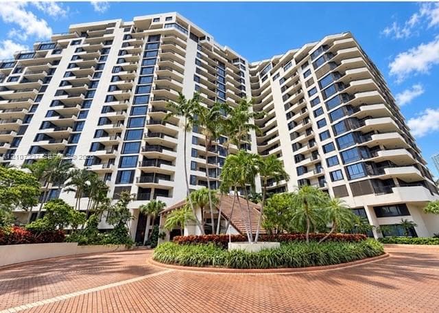 High-rise building exterior featuring tiered concrete balconies, floor-to-ceiling windows, a circular paved motor court, and landscaped gardens with mature palm trees