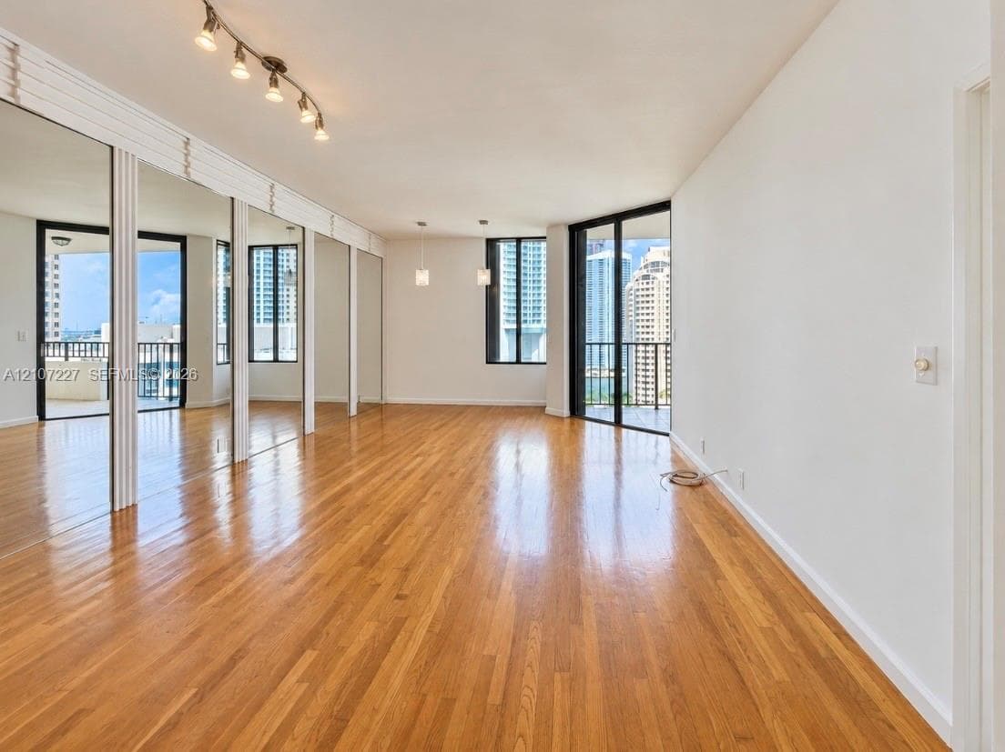 Open interior space featuring wood-finish flooring, floor-to-ceiling mirrored walls, sliding glass balcony doors, track lighting, and geometric multi-light fixtures