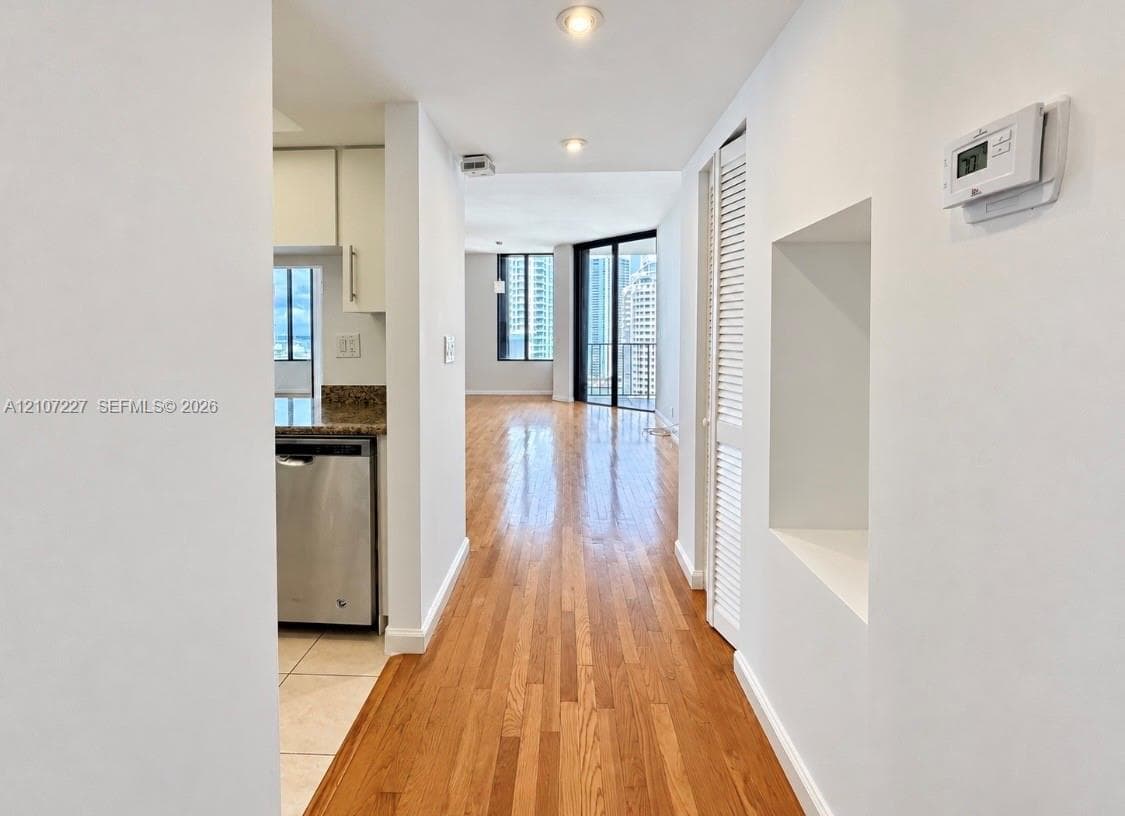 Hallway featuring wood-finish flooring, recessed lighting, and a built-in wall niche, with a partial view of a kitchen equipped with stainless steel dishwasher and stone-finish countertops