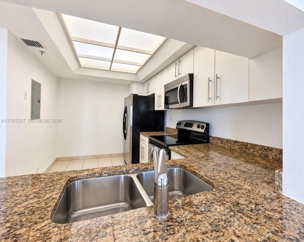 Kitchen featuring speckled stone countertops, dual-basin stainless steel sink, flat-panel white cabinetry, recessed tray ceiling lighting, and stainless steel appliances