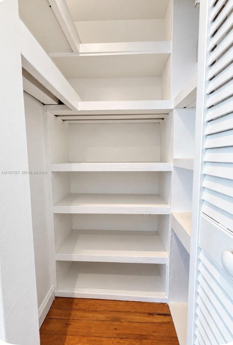 Storage space featuring built-in shelving, white-painted finish, and wood-finish flooring, accessed by a louvered door