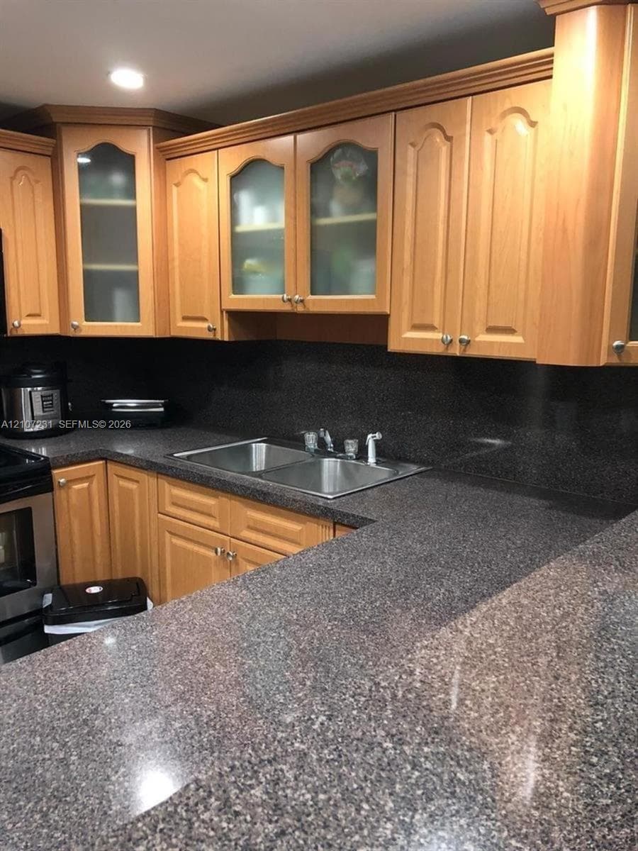 Kitchen featuring light wood-tone cabinetry, glass-paneled upper cabinet doors, dark solid-surface countertops, and a dual-basin stainless steel sink