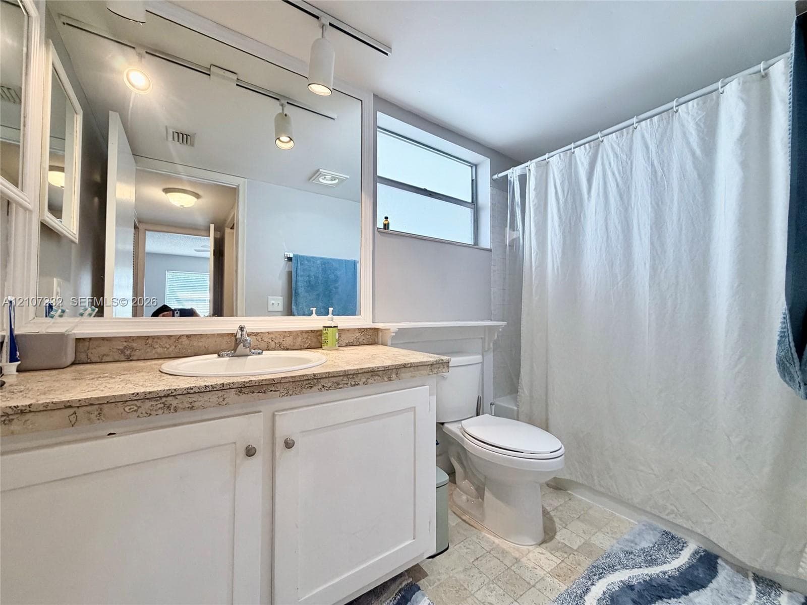 Bathroom featuring a stone-look countertop, integrated vanity, wall-to-wall mirror, track lighting, and a window above the toilet