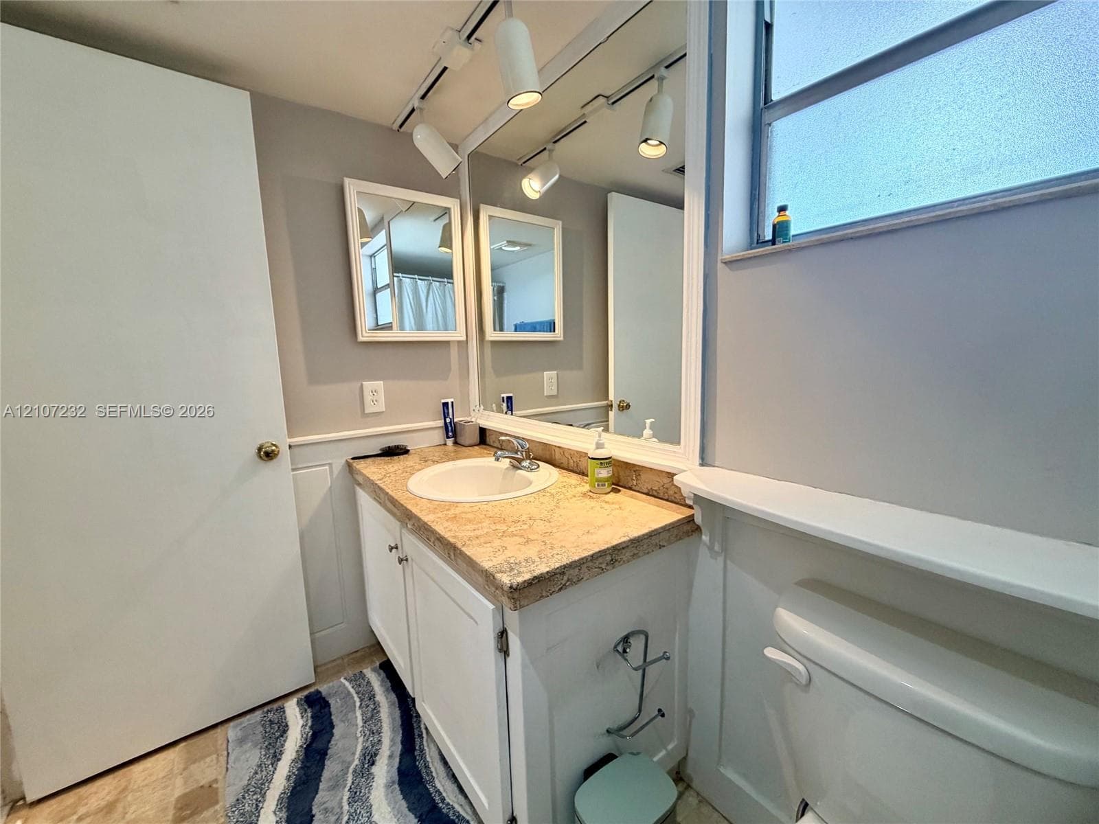 Vanity featuring a stone-look countertop with an integrated basin, white shaker-style cabinetry, wall-to-wall mirror, track lighting, and a window ledge