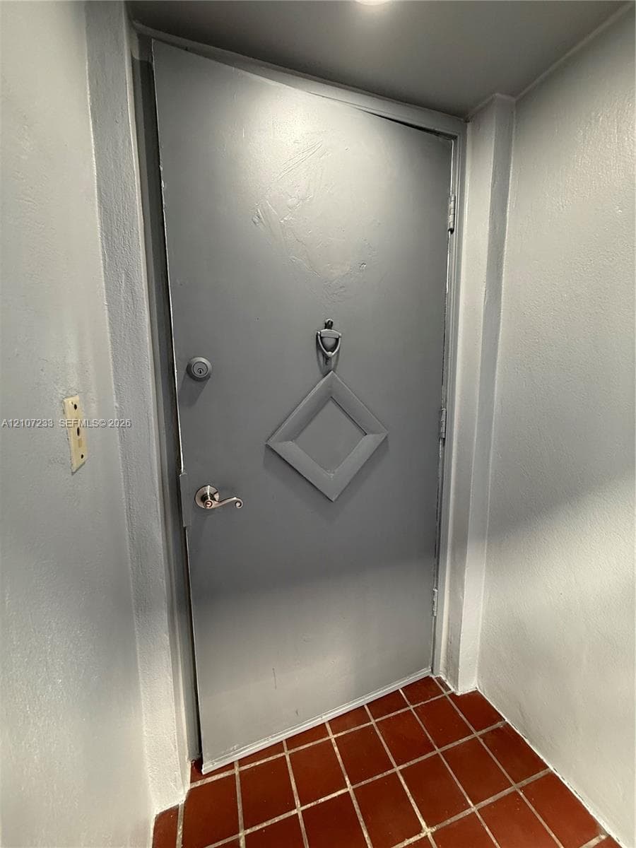 Solid door featuring a geometric diamond-shaped panel, chrome finish lever handle, and a decorative metal door knocker, set against red ceramic floor tiling