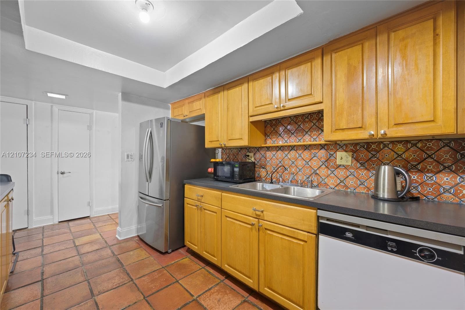 Kitchen featuring wood cabinetry, patterned tile backsplash, stainless steel refrigerator, dual-basin sink, and terracotta floor tiling