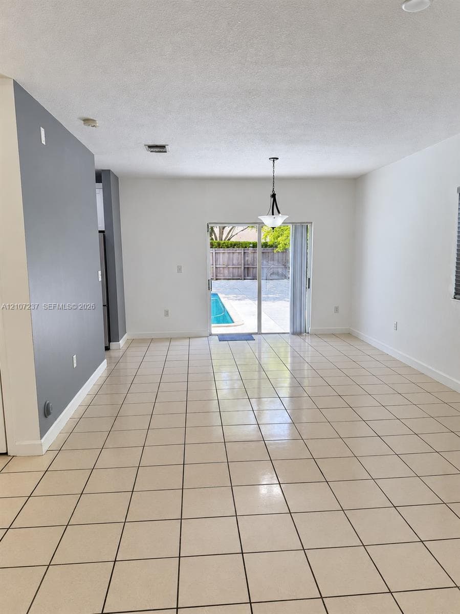 Neutral-tone interior featuring ceramic tile flooring, a dark-toned accent wall, a tiered hanging light fixture, and sliding glass doors opening to an exterior pool area