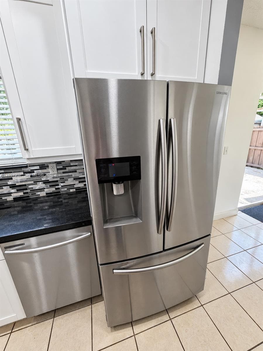 Stainless steel French door refrigerator, white shaker-style cabinetry, dark stone countertop, multi-tonal glass tile backsplash, and ceramic tile flooring