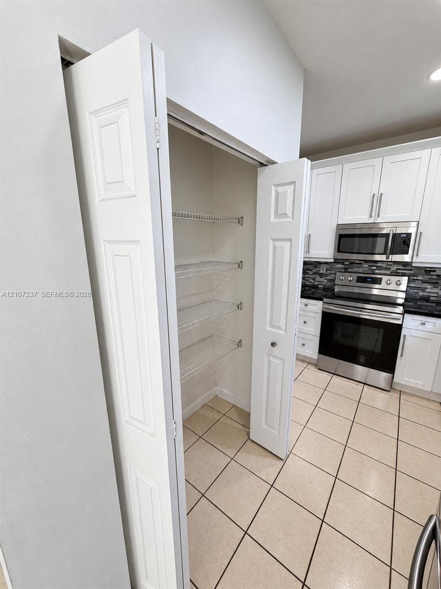 Pantry featuring wire shelving and bi-fold doors, complemented by kitchen cabinetry with stainless steel appliances, a multi-tonal backsplash, and ceramic tile flooring