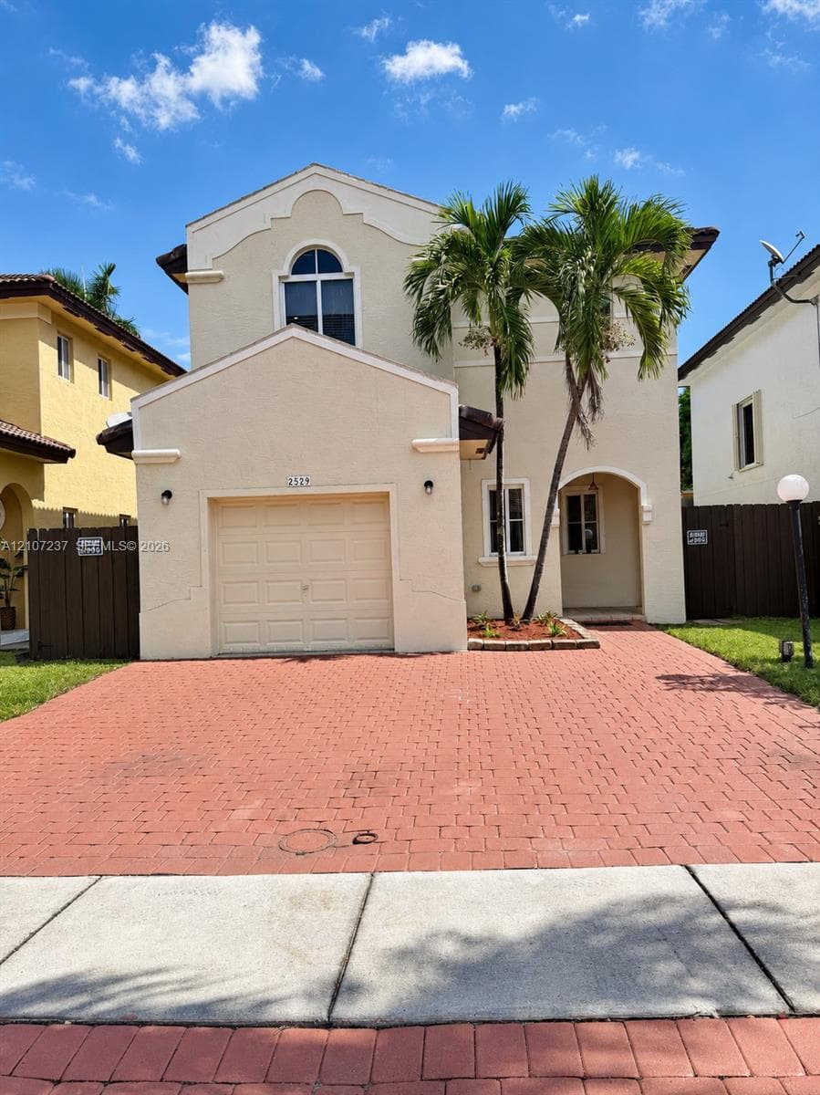 Two-story stucco exterior featuring a paved driveway, single-car garage, arched entryway, and a decorative upper-level window