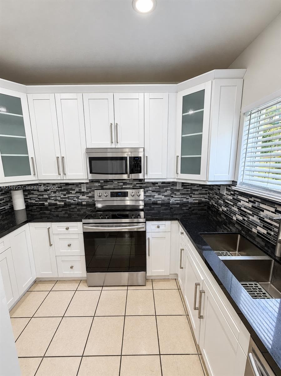 White shaker-style cabinetry with glass-front uppers, stainless steel appliances, dark stone countertops, mosaic tile backsplash, and dual-basin stainless steel sink