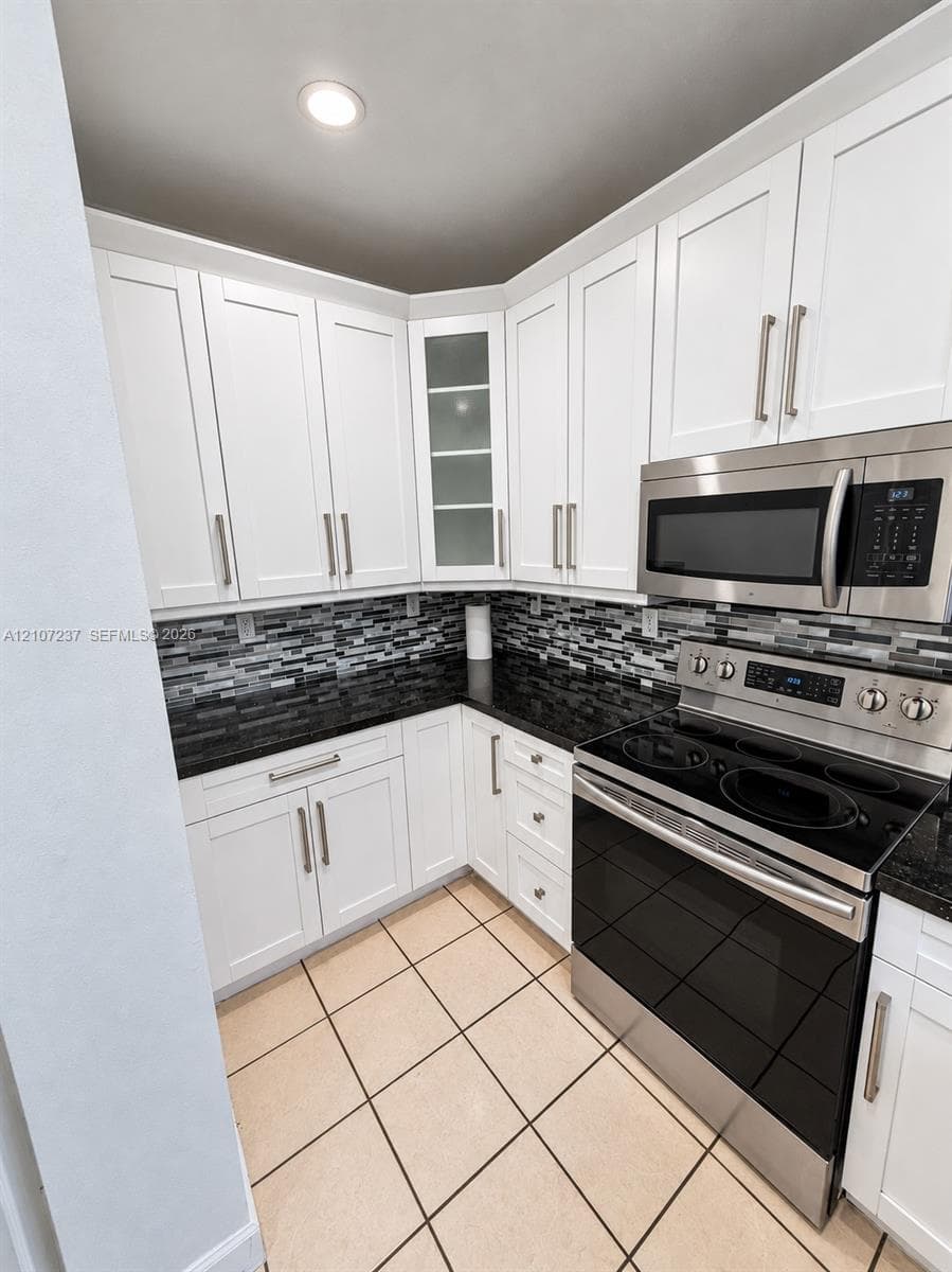 White shaker-style cabinetry with glass-front display accents, dark stone-finish countertops, mosaic tile backsplash, stainless steel appliances, and tile flooring