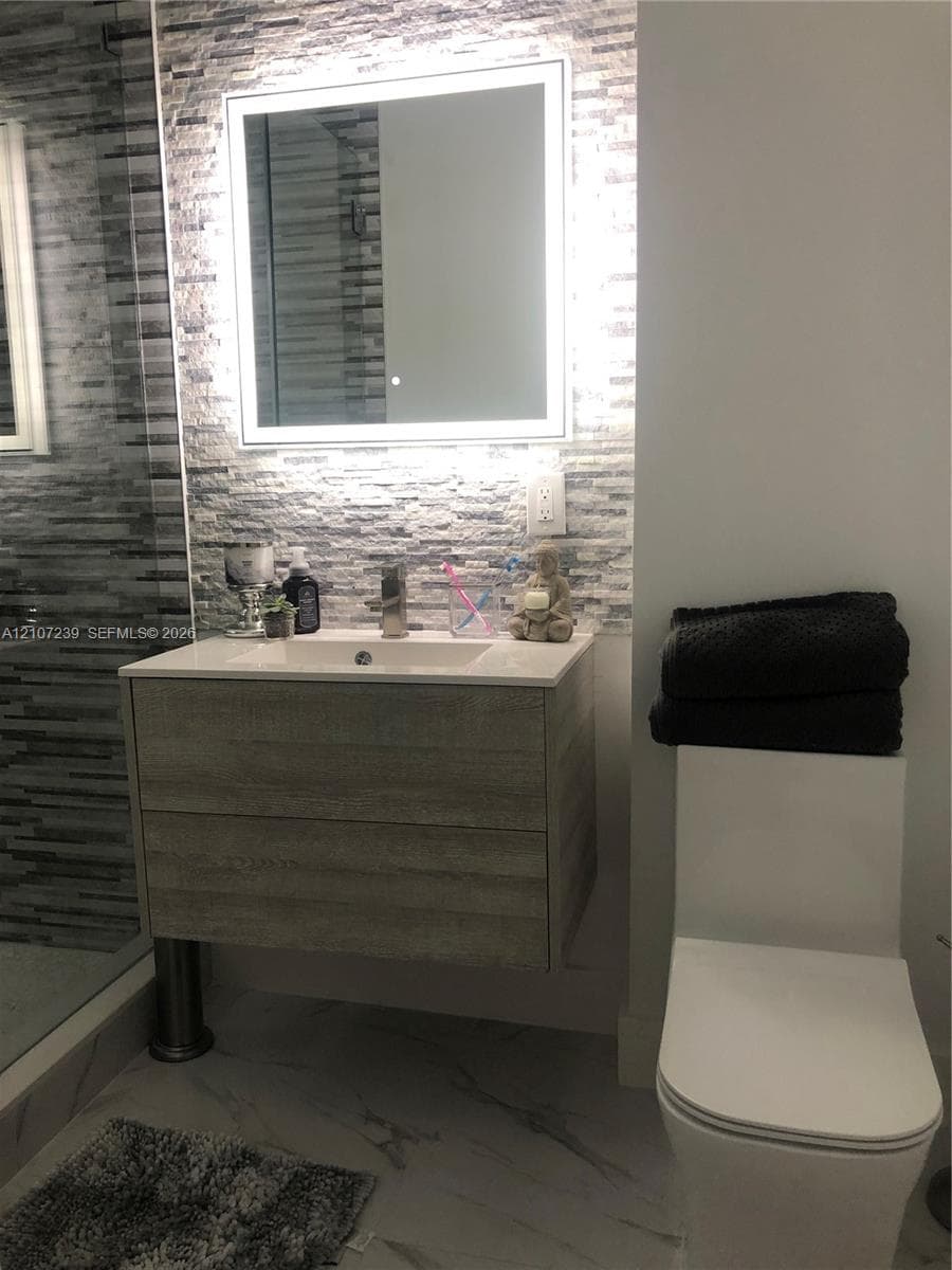 Floating vanity with wood-finish cabinetry, rectangular integrated sink, LED-backlit mirror, stacked stone-texture wall accent, and porcelain tile flooring