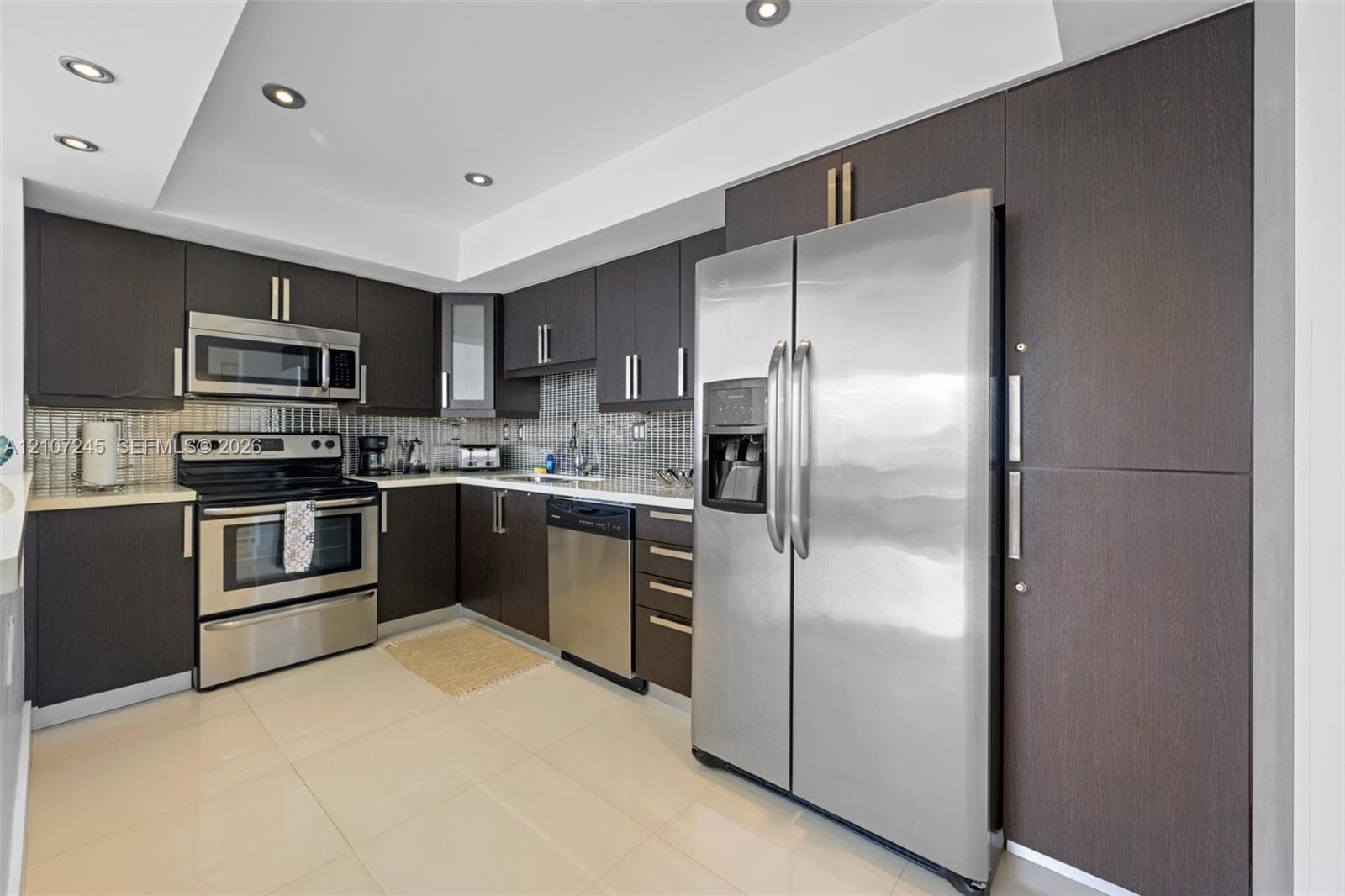Kitchen featuring dark-toned flat-panel cabinetry, stainless steel appliances, tiled backsplash, light-toned flooring, and recessed ceiling lighting
