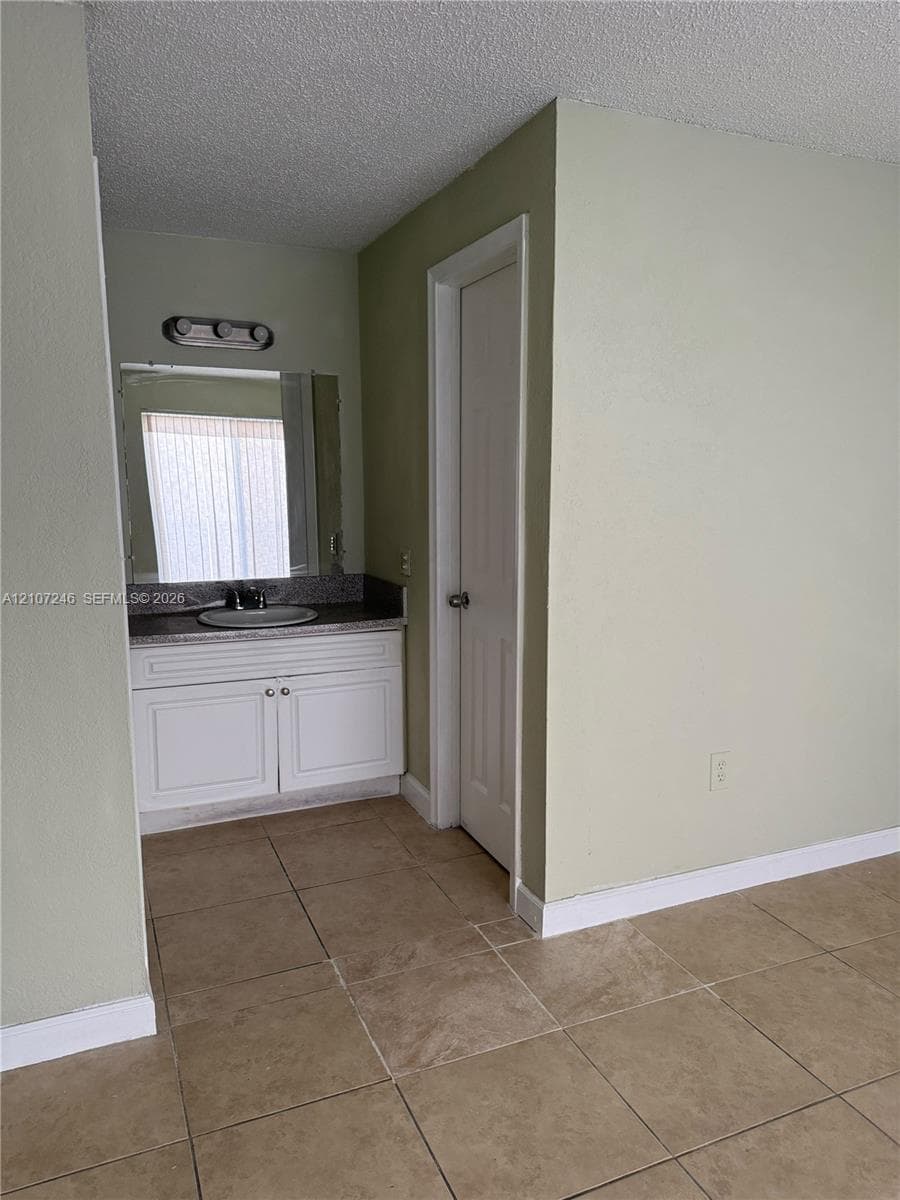 Vanity area featuring white cabinetry, stone-finish countertop with an integrated basin, wall-mounted mirror with integrated lighting, and tile flooring