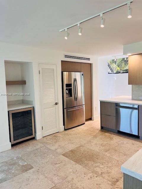 Kitchen featuring neutral stone flooring, wood-finish cabinetry, stainless steel appliances, built-in beverage refrigerator, and adjustable track lighting