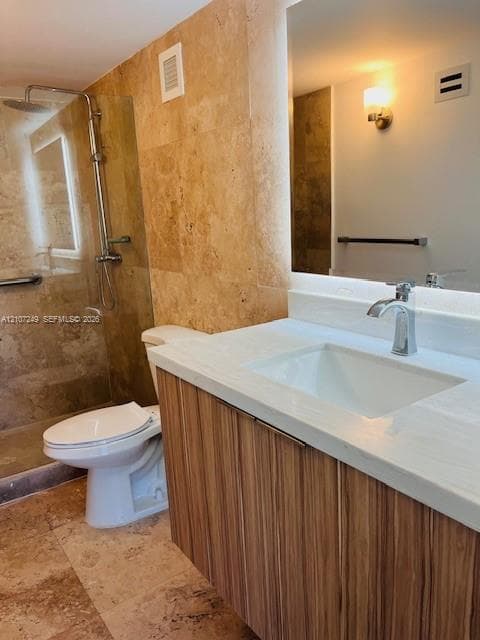 Floating wood-finish vanity with a white countertop and rectangular undermount sink, walk-in shower with a glass panel, stone-tiled wall and floor surfaces, and wall-mounted brushed metal faucet hardware
