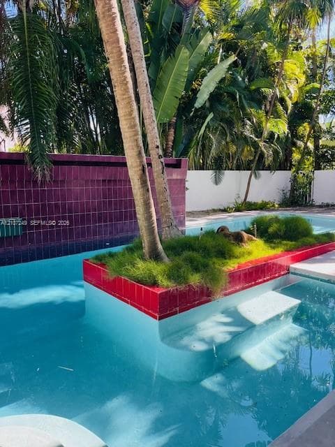 In-ground pool featuring a central concrete island with integrated landscaping, red ceramic tile accents, submerged entry steps, and a decorative purple tiled privacy wall