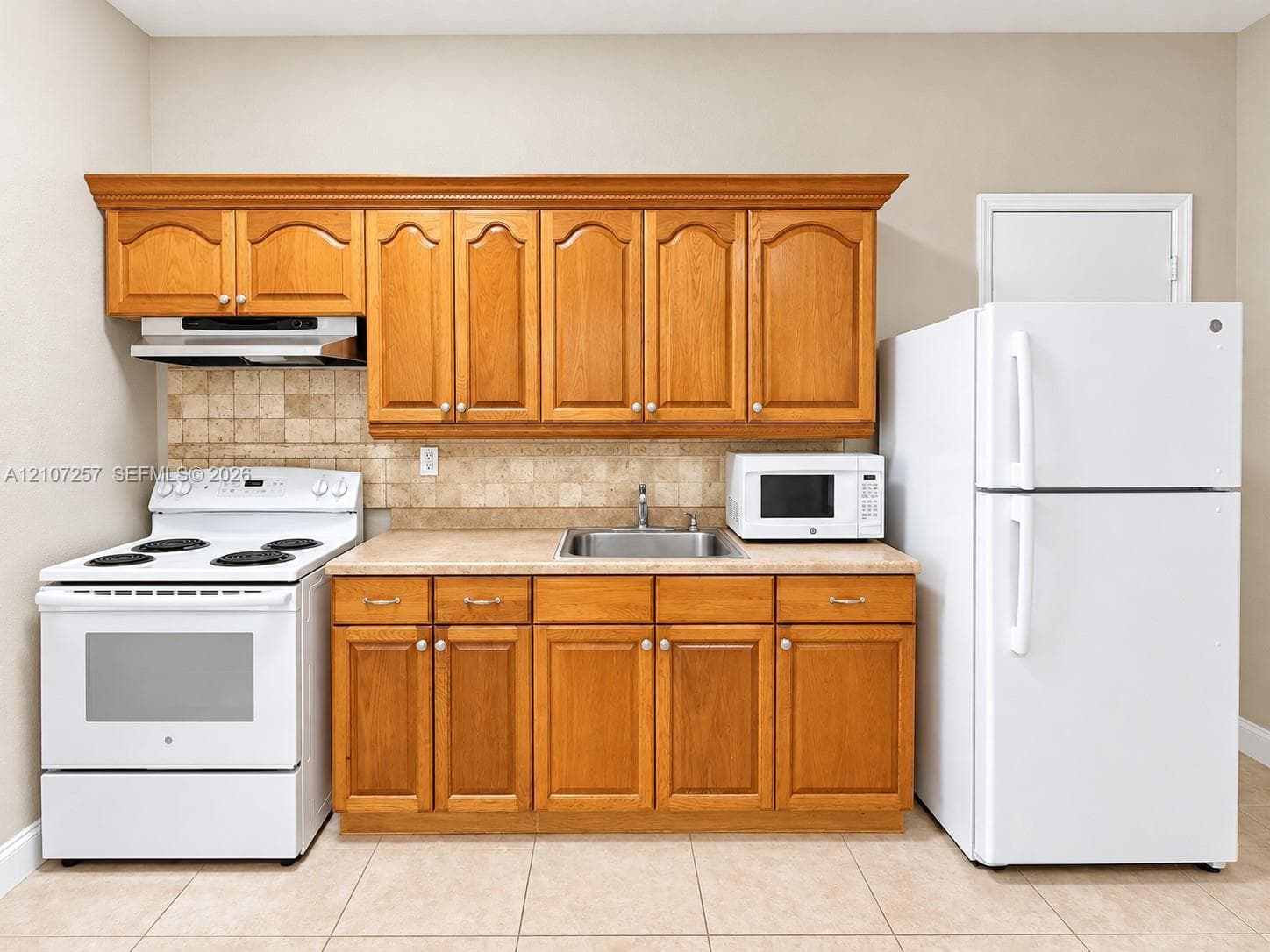 Kitchenette featuring natural wood cabinetry, light-toned stone-finish countertop, square-tile backsplash, stainless steel drop-in sink, and white appliances