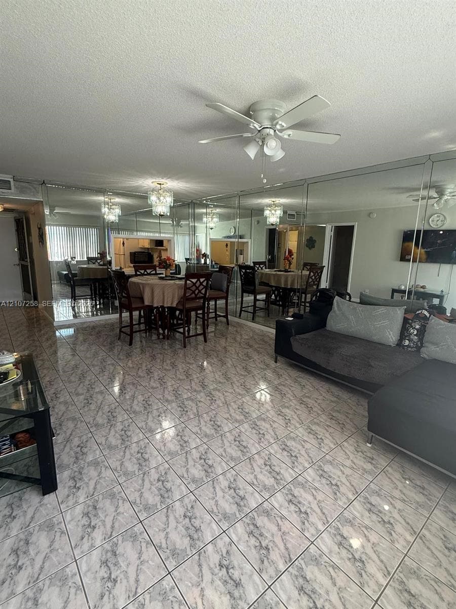 Spacious room featuring floor-to-ceiling mirrored walls, patterned tile flooring, a multi-blade ceiling fan, and a built-in crystal-style chandelier