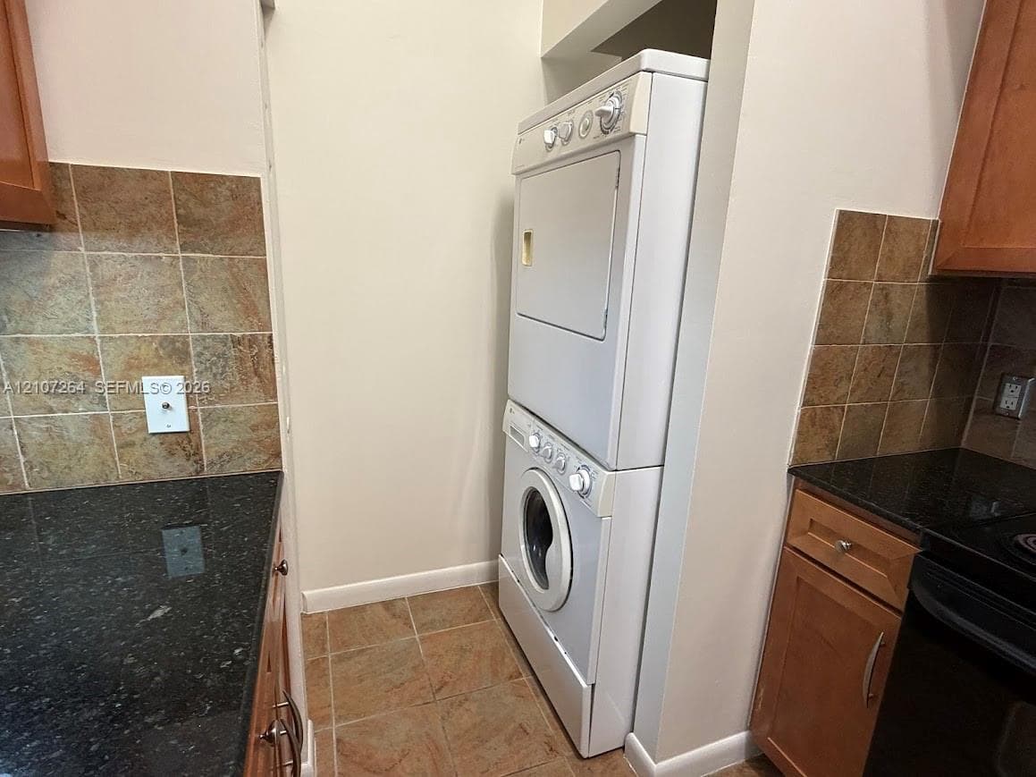 Stacked laundry unit enclosed by cabinetry, featuring stone-tile backsplash, dark solid-surface countertops, and wood-finish cabinetry