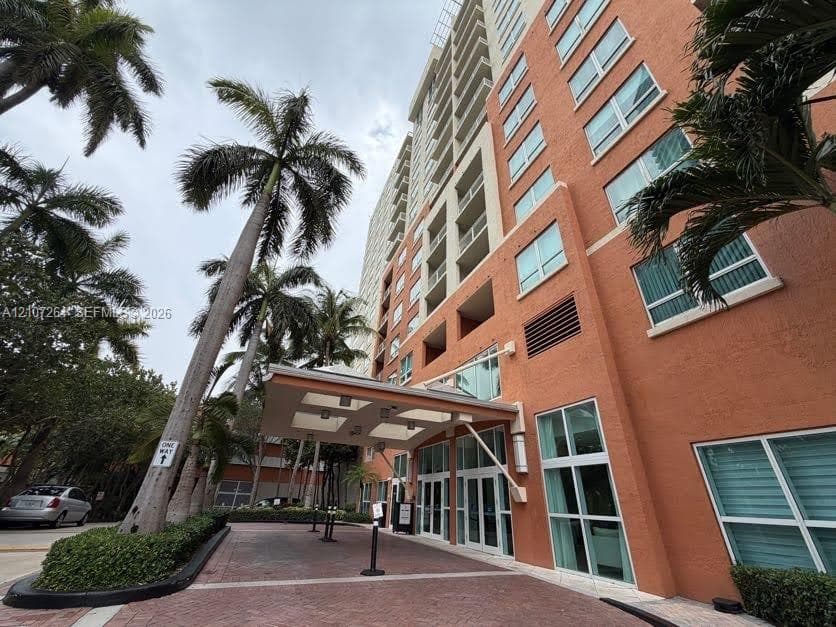 High-rise exterior featuring a structured porte-cochère, textured masonry facade, multi-story glass fenestration, and landscaped palm trees along a paver stone arrival court