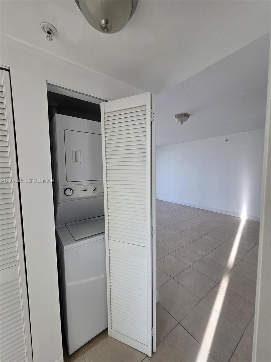 Laundry closet featuring a stackable washer and dryer, white louvered bi-fold doors, and tile flooring