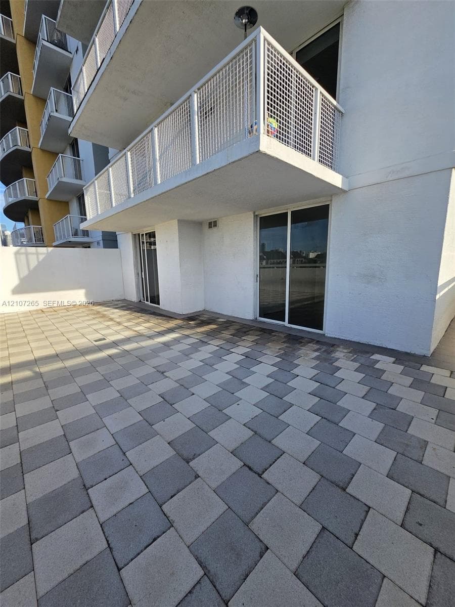 Expansive paved terrace featuring a multi-tone grid pattern, sliding glass doors, and a metal-railed balcony overhead