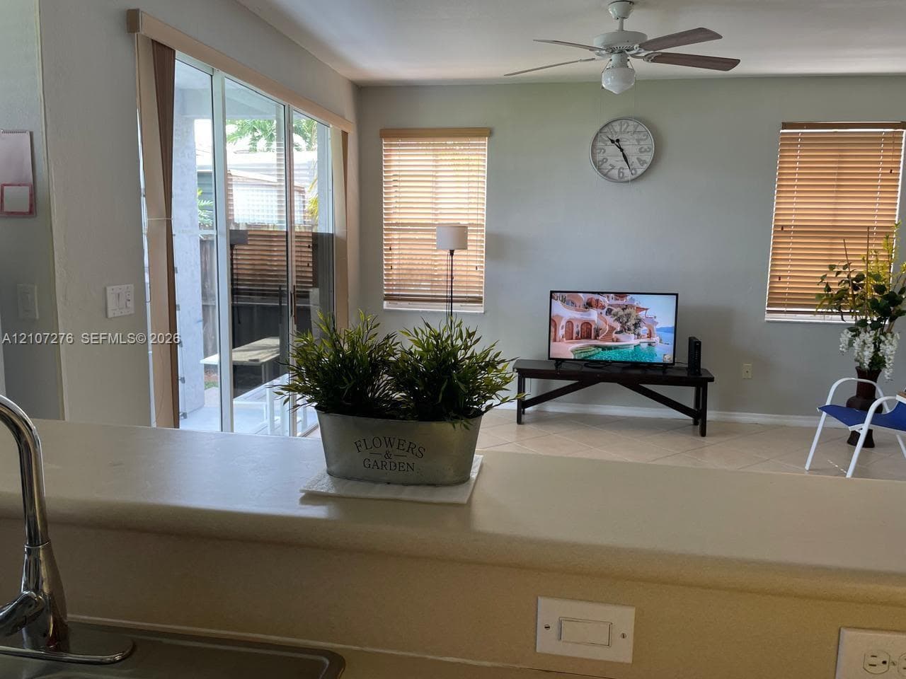 Neutral-toned living space featuring tile flooring, sliding glass patio doors, large windows with horizontal blinds, and a ceiling fan