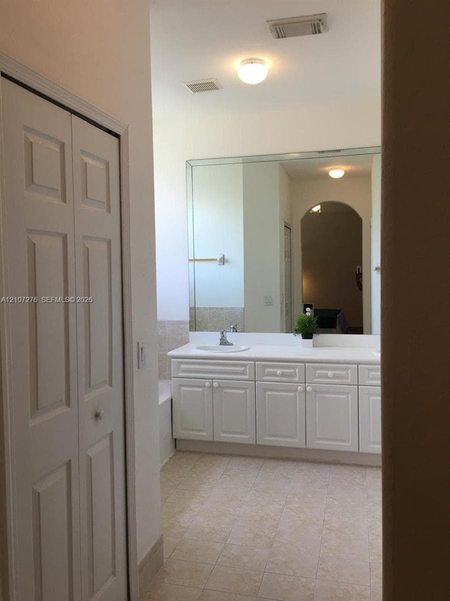 Bathroom featuring white shaker-style cabinetry, large wall-mounted mirror, stone-finish tile flooring, and integrated white countertop with a recessed sink