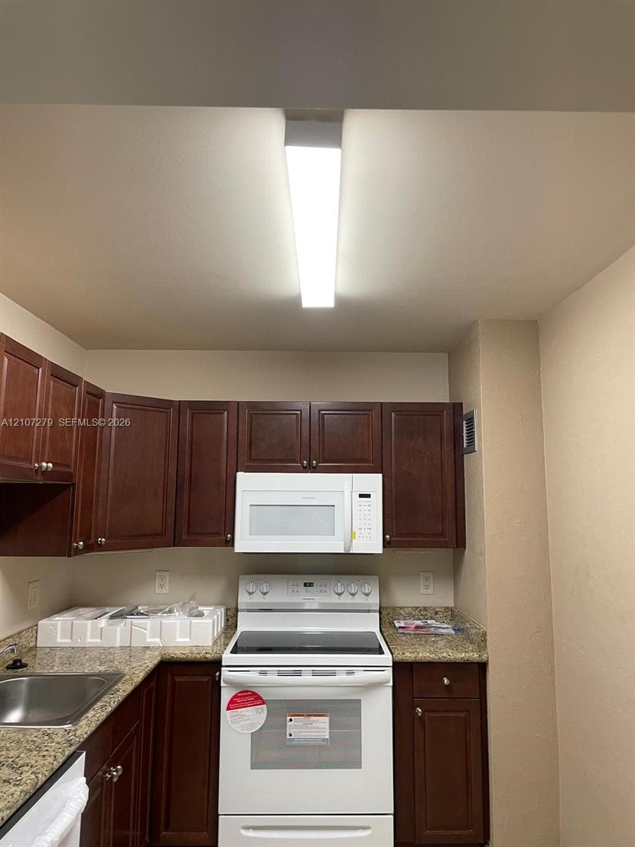 Kitchen featuring dark wood cabinetry, stone-finish countertops, stainless steel sink, and white appliances including a built-in microwave and range