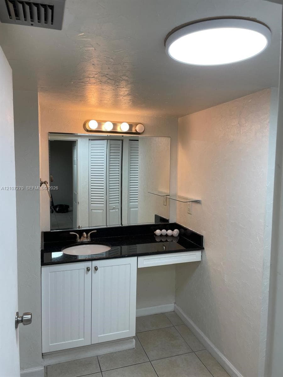 Vanity area featuring white shaker-style cabinetry, dark stone-finish countertop with an integrated basin, large rectangular mirror, and dual ceiling-mounted lighting fixtures