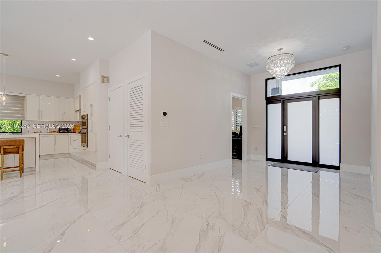 Large-format polished tile flooring, glass-panel front door with transom window, high-gloss white cabinetry, integrated wall oven, and tiered crystal chandelier