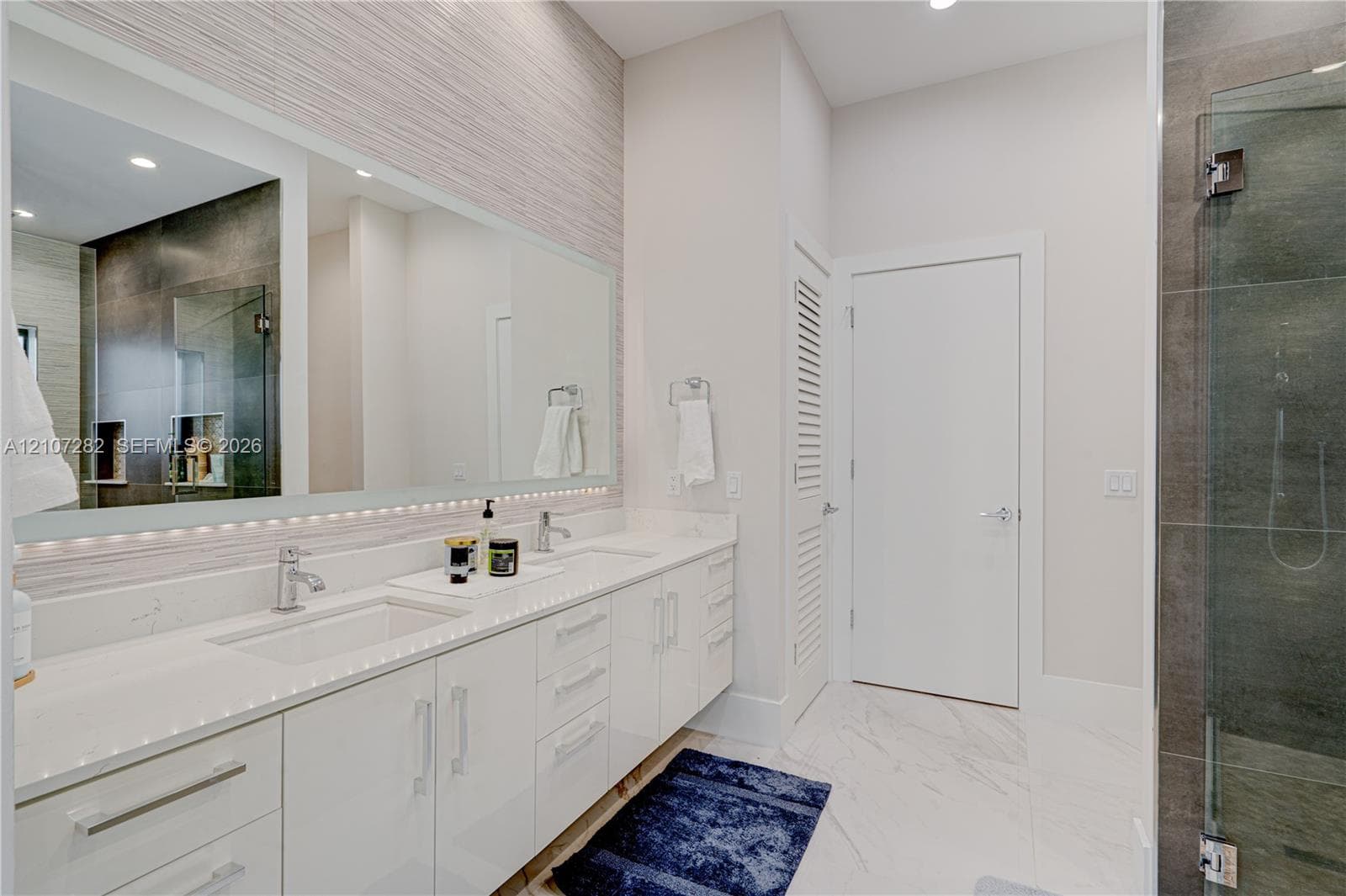 Double vanity with white cabinetry, light-toned stone-finish countertops, undermount sinks, large wall-spanning mirror, and a walk-in glass-enclosed shower with tiled walls