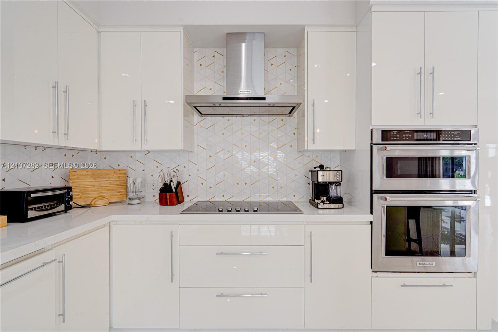 Kitchen featuring high-gloss white cabinetry, geometric patterned backsplash, stainless steel double wall oven, stainless steel range hood, and solid-surface countertops